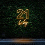 Yellow neon sign reading "21 Baby," designed for 21st birthday party decorations, glowing against a lush green leafy wall backdrop.