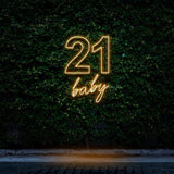Yellow neon sign reading "21 Baby," designed for 21st birthday party decorations, glowing against a lush green leafy wall backdrop.