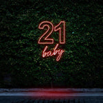 Bright red neon sign reading "21 Baby," glowing against a green leafy wall, perfect for 21st birthday celebrations and bold party decorations.