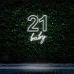 Bright white neon sign reading "21 Baby," glowing elegantly against a green leafy wall, ideal for 21st birthday celebrations and minimalist party decorations.