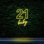 Bright yellow neon sign reading "21 Baby," glowing against a green leafy wall, ideal for 21st birthday celebrations and festive party decorations.

