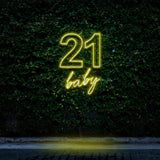 Bright yellow neon sign reading "21 Baby," glowing against a green leafy wall, ideal for 21st birthday celebrations and festive party decorations.

