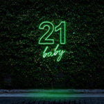 Neon sign reading "21 Baby" in bright green light, perfect for 21st birthday celebrations and party decorations, displayed against a leafy wall background.