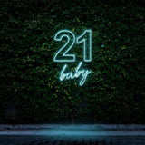 Illuminated "21 Baby" neon sign in vibrant blue, perfect for 21st birthday party decorations, featuring a glowing design on a lush green background for stylish celebrations.