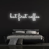 Cold white "But First Coffee" LED neon sign displayed above a sophisticated bed setup with a dark wall background, ideal for minimalist coffee enthusiasts.