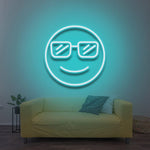 A smiling cool emoji with sunglasses, glowing in aqua blue LED neon light, mounted on a wall above a yellow sofa in a modern living room with a cozy vibe.

