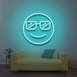 A smiling cool emoji with sunglasses, glowing in aqua blue LED neon light, mounted on a wall above a yellow sofa in a modern living room with a cozy vibe.

