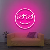 A smiling cool emoji with sunglasses, glowing in pink LED neon light, mounted on a wall above a yellow sofa in a cozy living room with modern decor.

