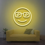 A smiling cool emoji with sunglasses, glowing in yellow LED neon light, mounted on a wall above a yellow sofa in a cozy living room with modern decor.

