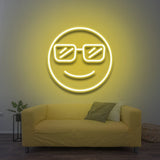 A smiling cool emoji with sunglasses, glowing in yellow LED neon light, mounted on a wall above a yellow sofa in a cozy living room with modern decor.

