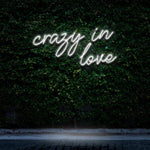 "Crazy In Love" LED Neon Sign glowing in cold white light, set against a lush green wall, offering a sleek and modern look as romantic wall decor.

