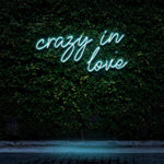 "Crazy In Love" LED Neon Sign glowing in green, set against a lush green wall, creating a romantic and modern ambiance for stylish wall decor.

