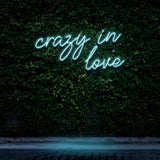"Crazy In Love" LED Neon Sign glowing in green, set against a lush green wall, creating a romantic and modern ambiance for stylish wall decor.

