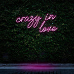 "Crazy In Love" LED Neon Sign glowing in pink, set against a lush green wall, creating a vibrant and romantic atmosphere for modern room decor.

