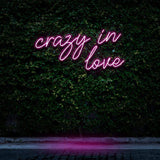 "Crazy In Love" LED Neon Sign glowing in pink, set against a lush green wall, creating a vibrant and romantic atmosphere for modern room decor.

