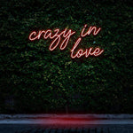 "Crazy In Love" LED Neon Sign glowing in red, set against a lush green wall, creating a bold and romantic atmosphere as modern wall decor.