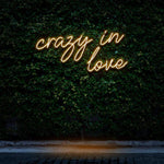 "Crazy In Love" LED Neon Sign glowing in warm white light, set against a green wall, adding a cozy and romantic vibe to the space as wall decor.