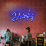 Blue "Drinks" LED neon sign illuminating a cozy cafe with a pink tiled wall.