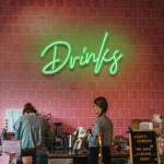 Green "Drinks" LED neon sign hanging in a trendy cafe with a pink-tiled backdrop.