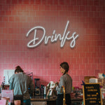 White "Drinks" LED neon sign featured in a stylish cafe with pink tiles and baristas at work.