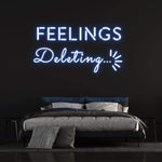 Blue "Feelings Deleting" neon sign on a dark wall above a modern bedroom setup, perfect for trendy home decor and wall art.