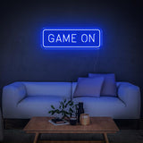 Blue GAME ON LED neon sign glowing above a white couch with pillows and wooden table.