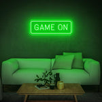 Green GAME ON LED neon sign glowing above a white couch with a wooden table and plants.