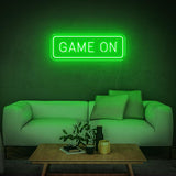 Green GAME ON LED neon sign glowing above a white couch with a wooden table and plants.