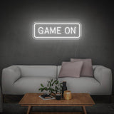 White GAME ON LED neon sign on the wall above a minimalist white couch with decor and table.
